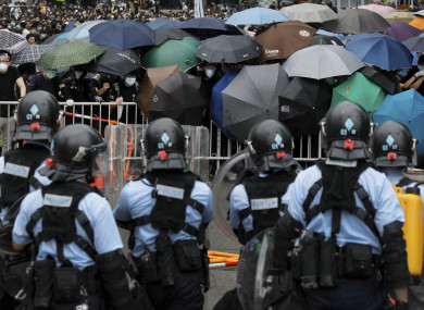Policemen in anti-riot gear stand watch as protesters use umbrellas to shield themselves.